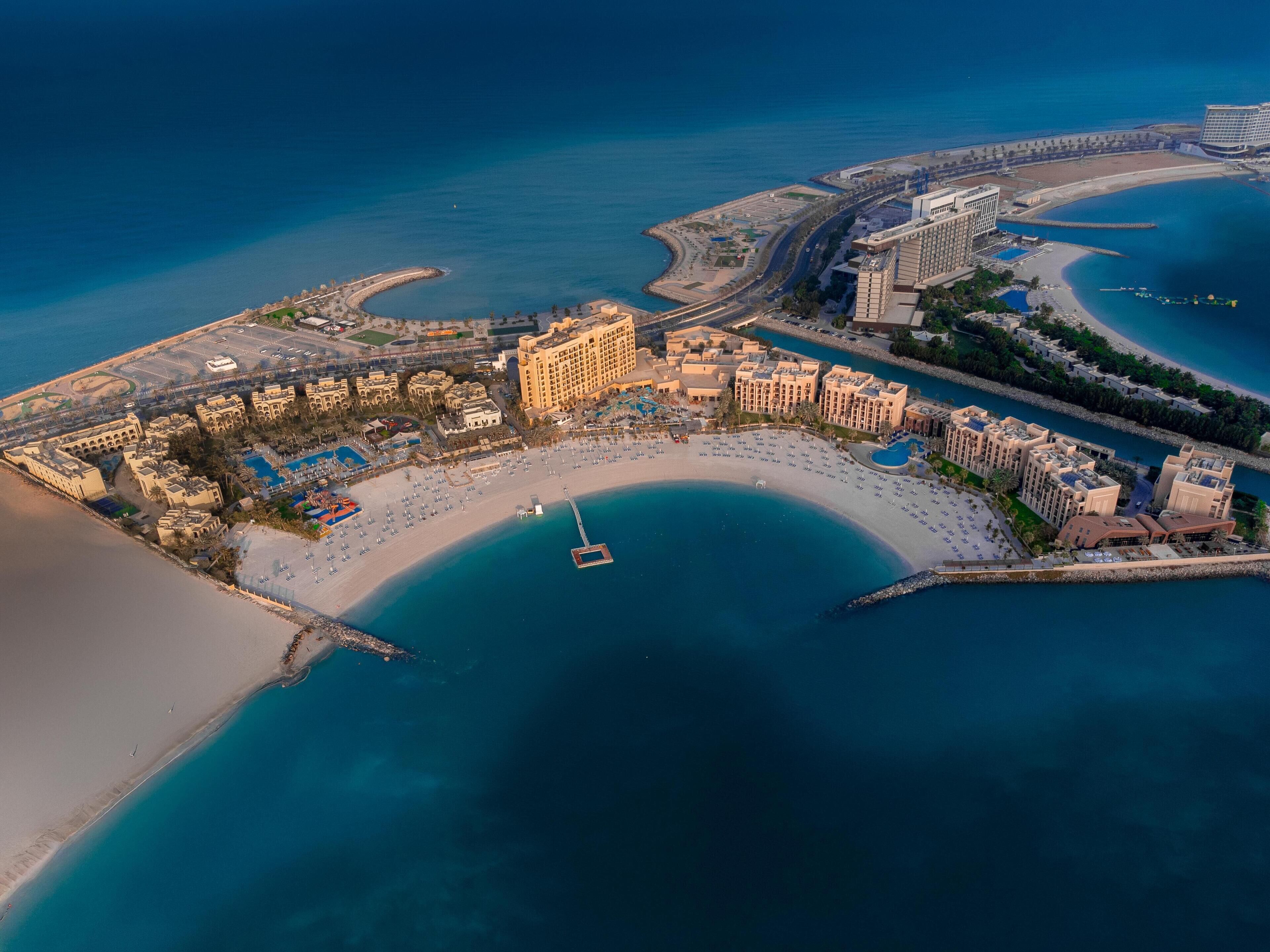 Vista aérea de un complejo turístico costero con villas frente a la playa, un puerto deportivo y un gran hotel.