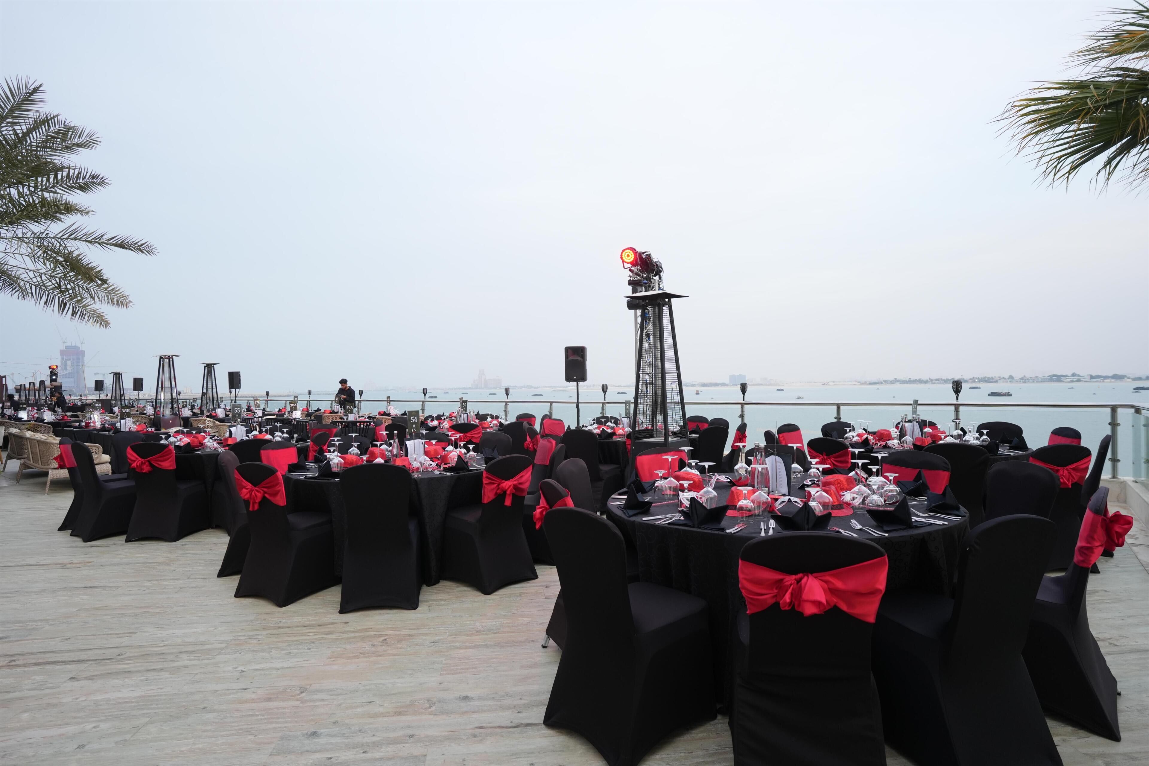 Un área de comedor al aire libre con mesas preparadas para los comensales, sillas con cintas rojas y vistas al mar.
