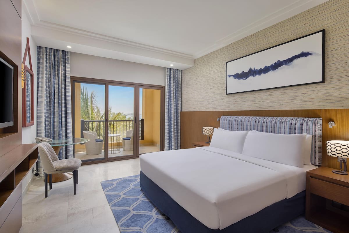 A hotel room with a bed, a TV, a desk, a chair, a balcony with a view, and a painting on the wall.