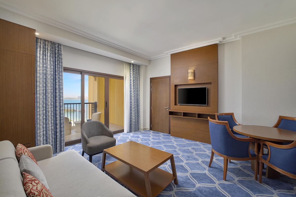 A hotel room with a balcony, a TV, a sofa, a coffee table, and a dining area.