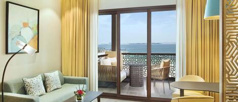 A hotel room with a sofa, a coffee table, a lamp, a chair, and a view of the sea through a large sliding door.