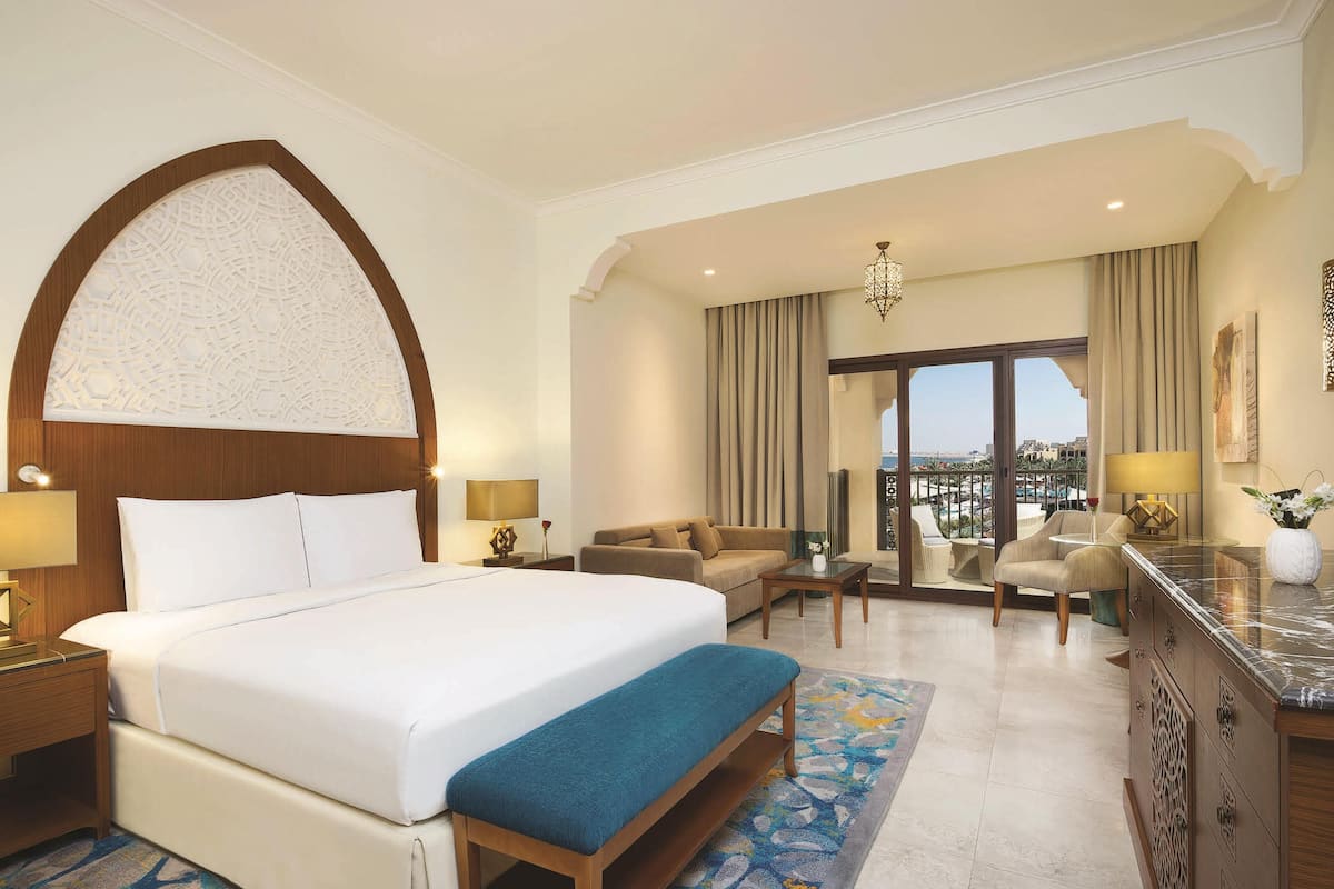 A hotel room with a large bed, a seating area with a sofa and chairs, a desk, and a view of the city.