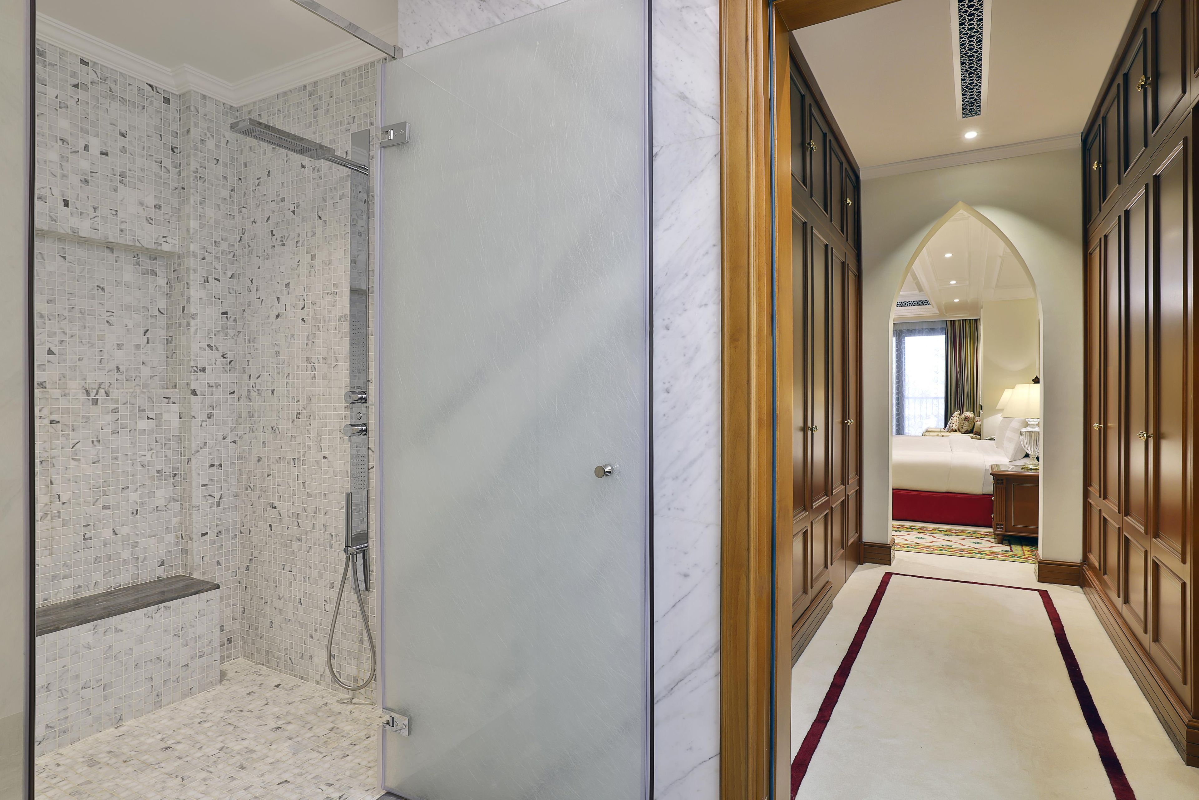 A hotel room with a marble shower, a wooden wardrobe, and a view of the adjacent bedroom.