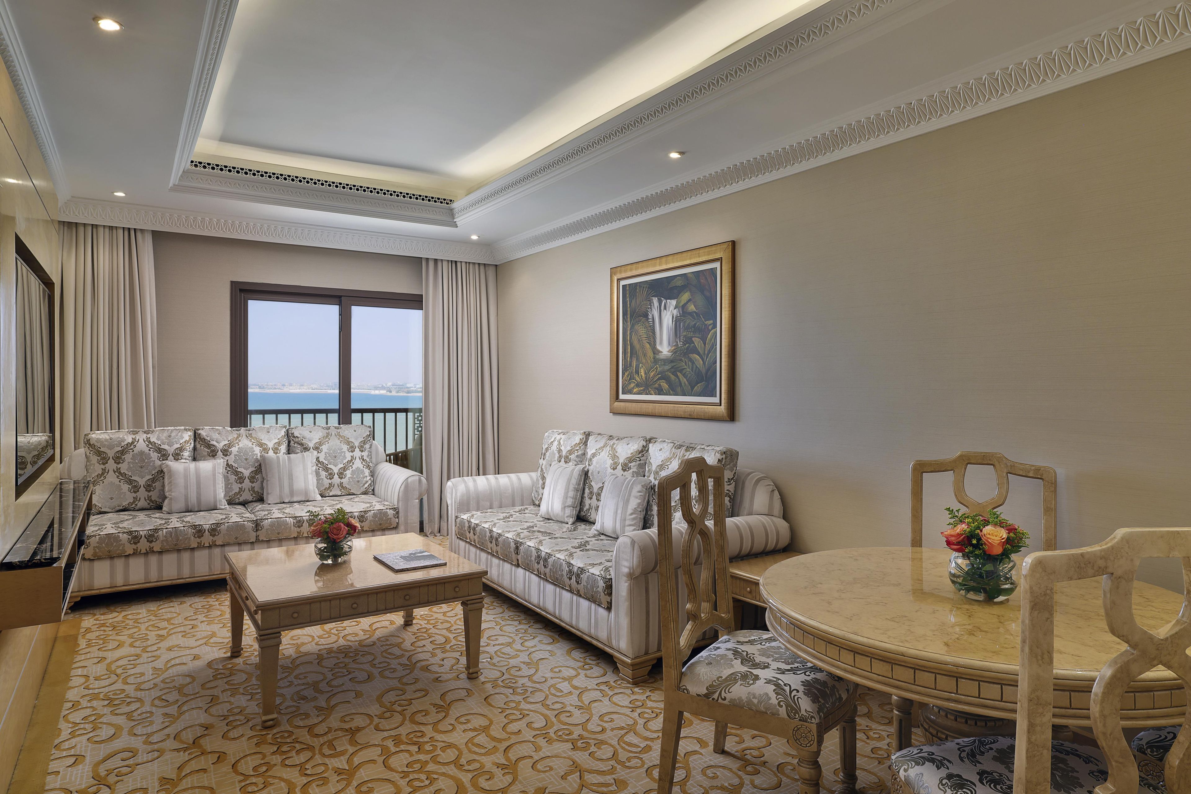 A hotel room with two sofas, a coffee table, a dining table with chairs, a painting on the wall, and a view of the sea through the window.