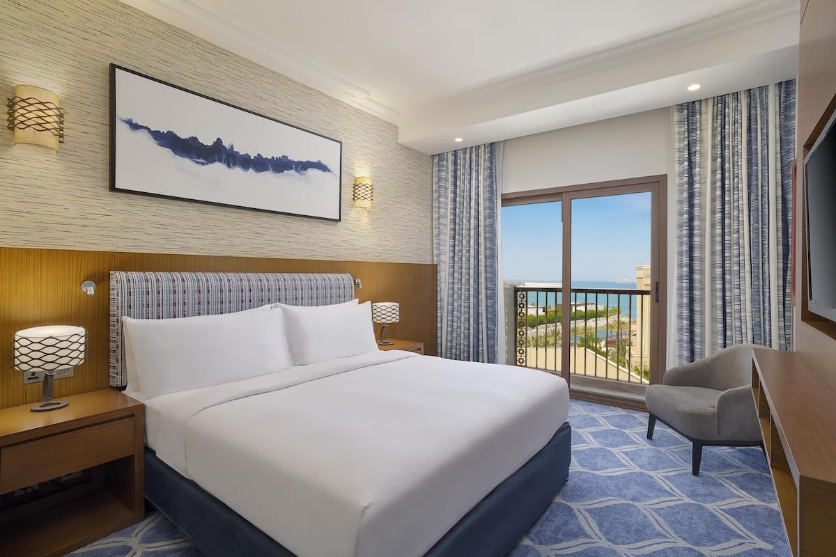 A hotel room with a large bed, a desk, a TV, a balcony with a view, and artwork on the wall.