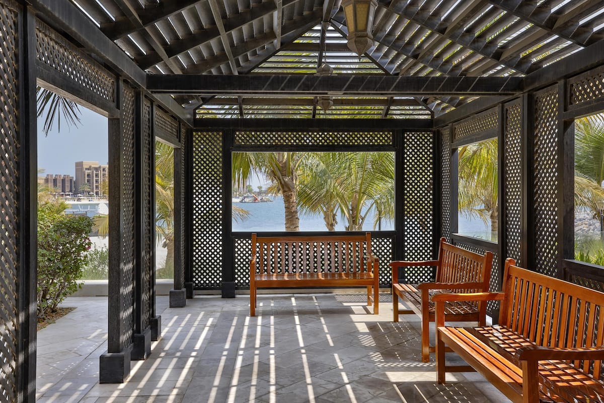A covered outdoor seating area with wooden benches and a view of palm trees and a body of water.