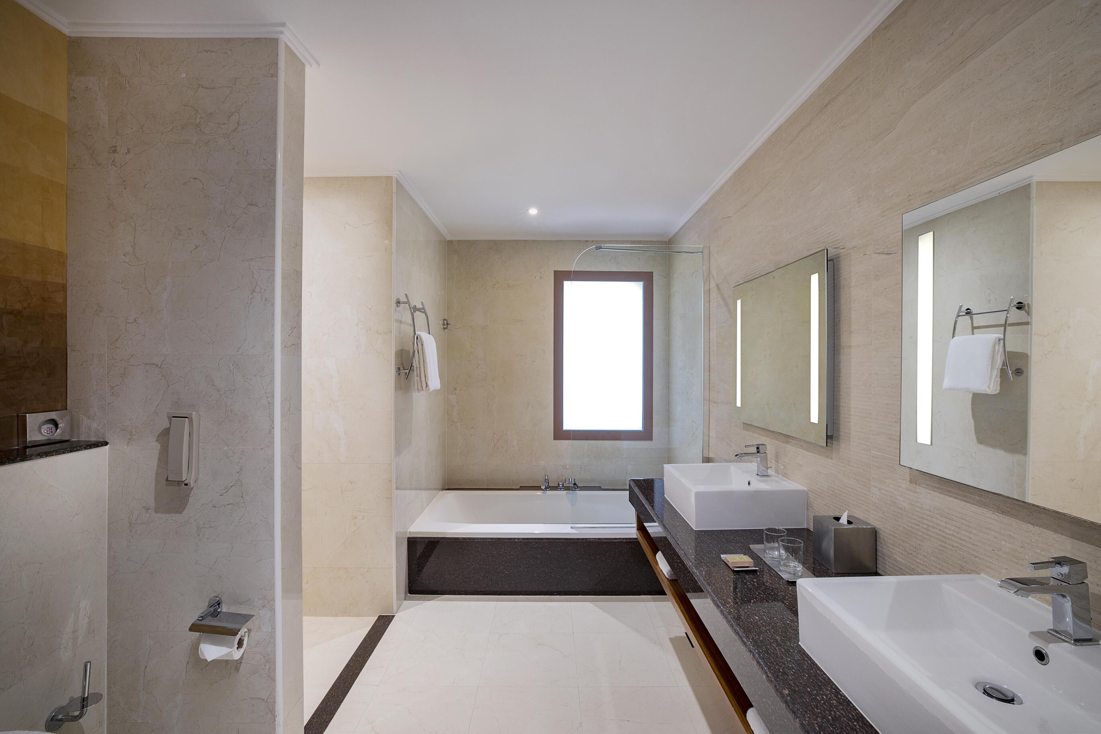 Een moderne badkamer met een badkuip, twee wastafels, een grote spiegel en een raam.