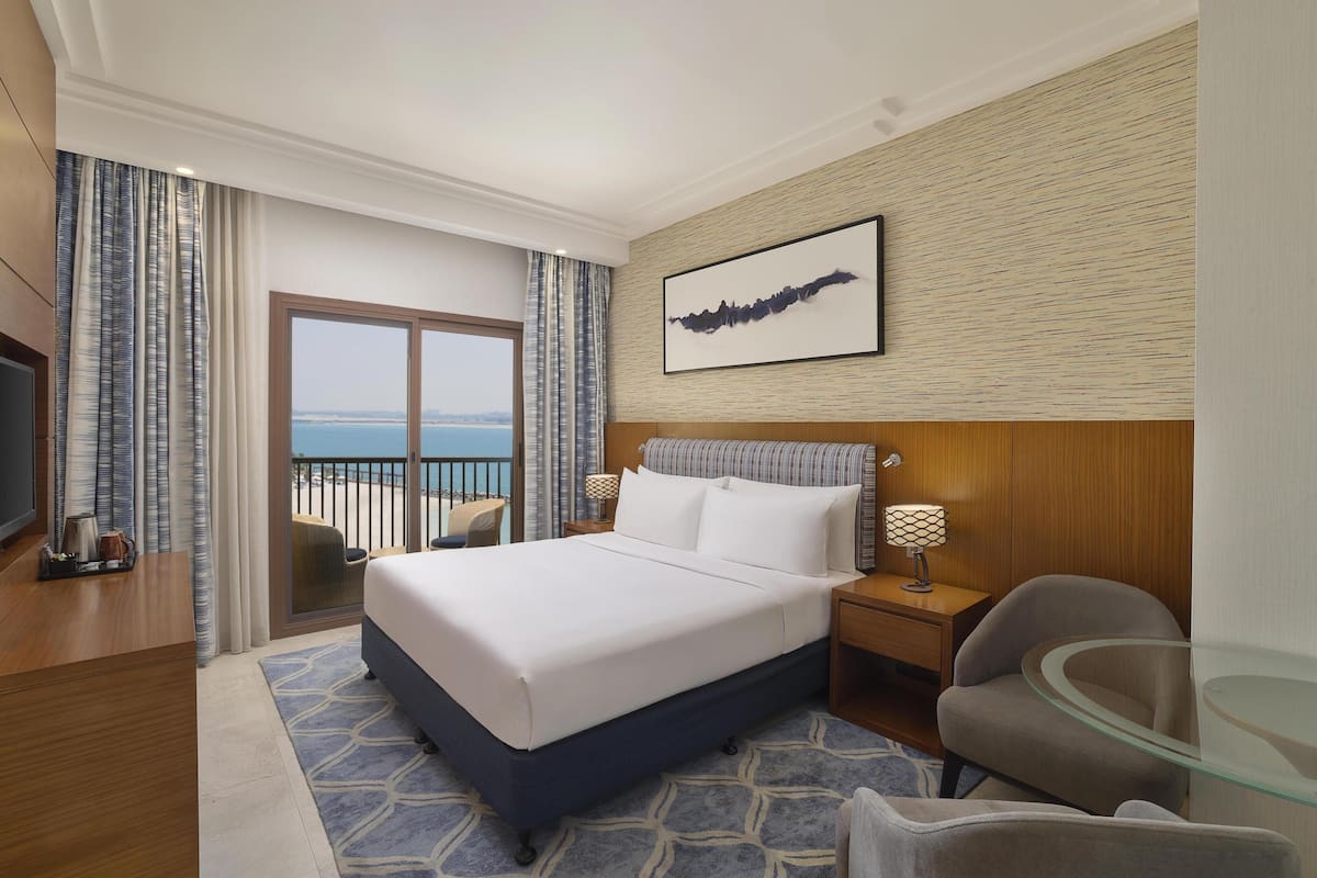 A hotel room with a large bed, a glass table, a chair, a TV, and a view of the sea.