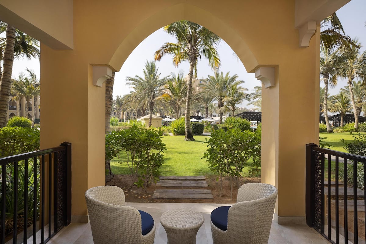 A covered outdoor seating area with wicker chairs and a view of a well-maintained garden with palm trees and a building in the background.