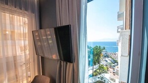 A hotel room with a large window, a flat-screen TV mounted on the wall, and a view of a coastal area with buildings and greenery. - Apollon Hotel (SARANDE)