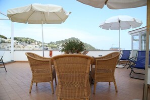 A terrace with wicker chairs and white umbrellas, overlooking a body of water and a hillside. - Guest House La Piazzetta (Capri)