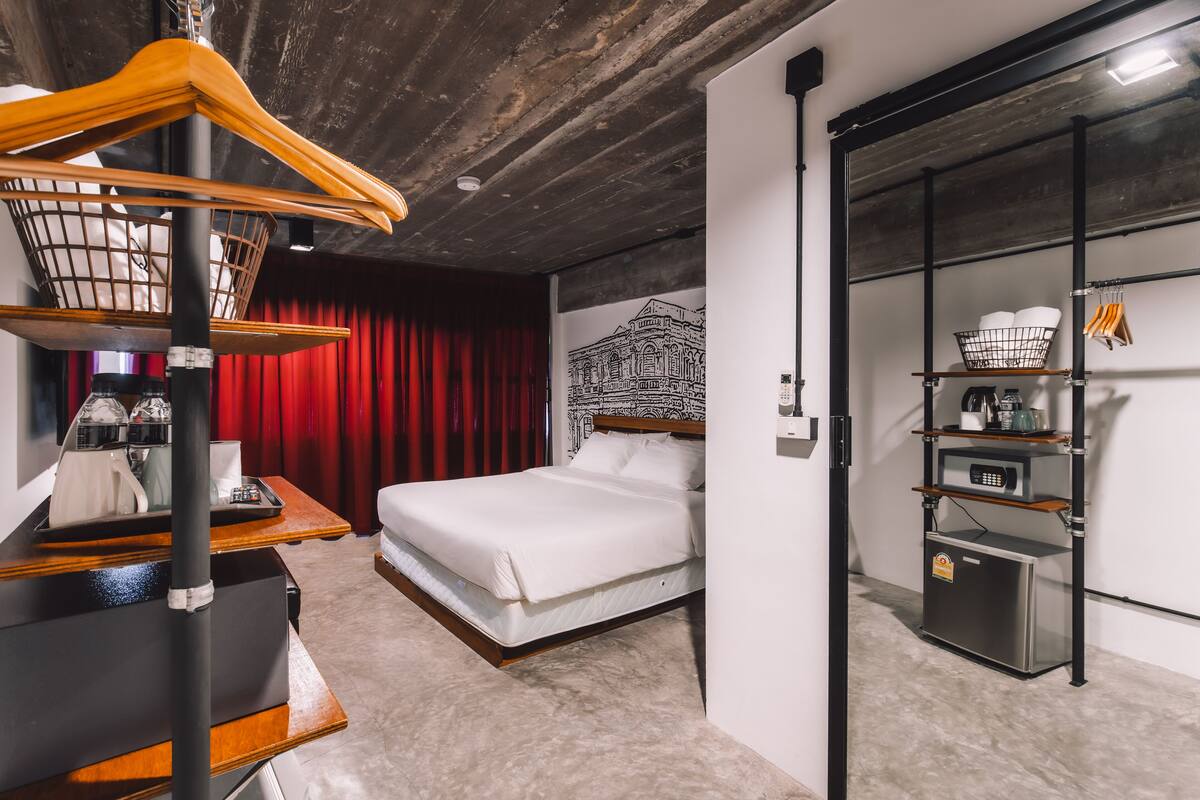 Ein Hotelzimmer mit einem Bett, einem Holzsessel, einem kleinen Tisch mit einem Korb, einem roten Vorhang, einer Wand mit einem Schwarz-Weiß-Muster und einer Küchenzeile mit Regalen.