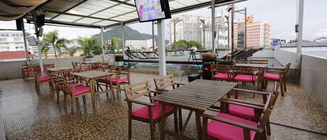 An outdoor dining area with wooden tables and chairs, a TV screen, and a view of buildings and hills in the background.