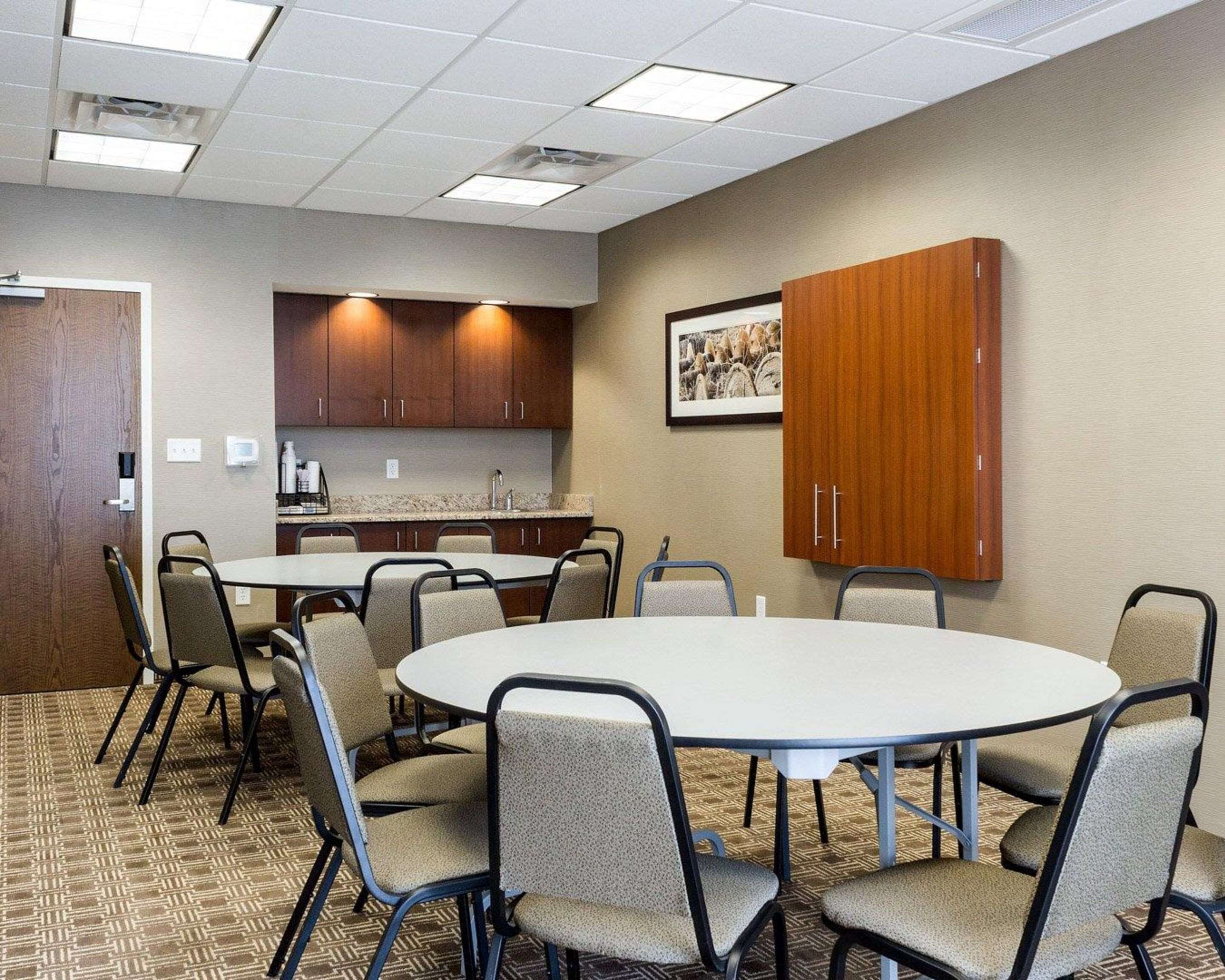Una sala de conferencias con una mesa redonda y ocho sillas, una kitchenette con armarios de madera, y un cuadro en la pared.