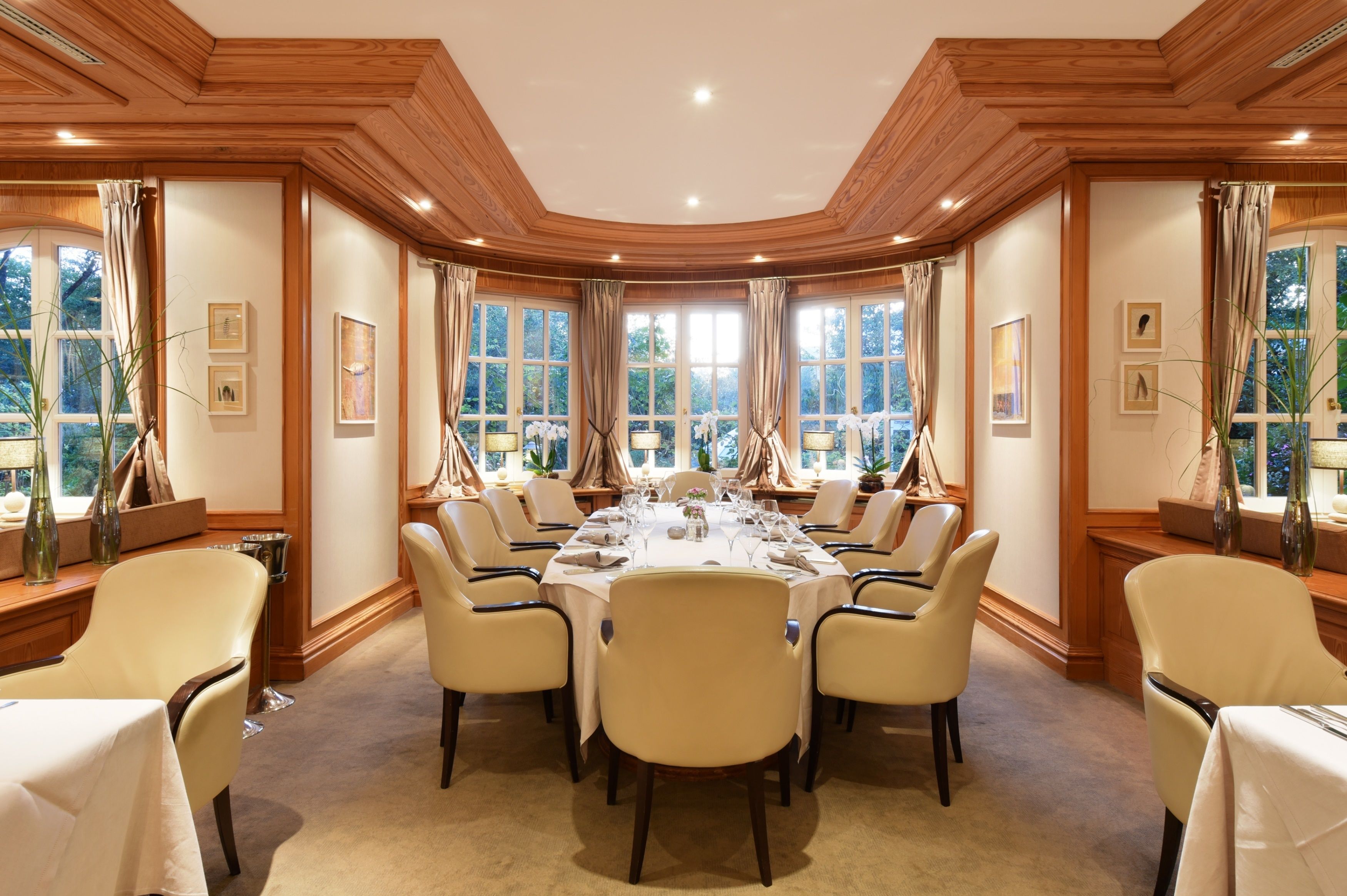 A formal dining room with a long table set for a meal, surrounded by chairs, with large windows and framed artwork on the walls.