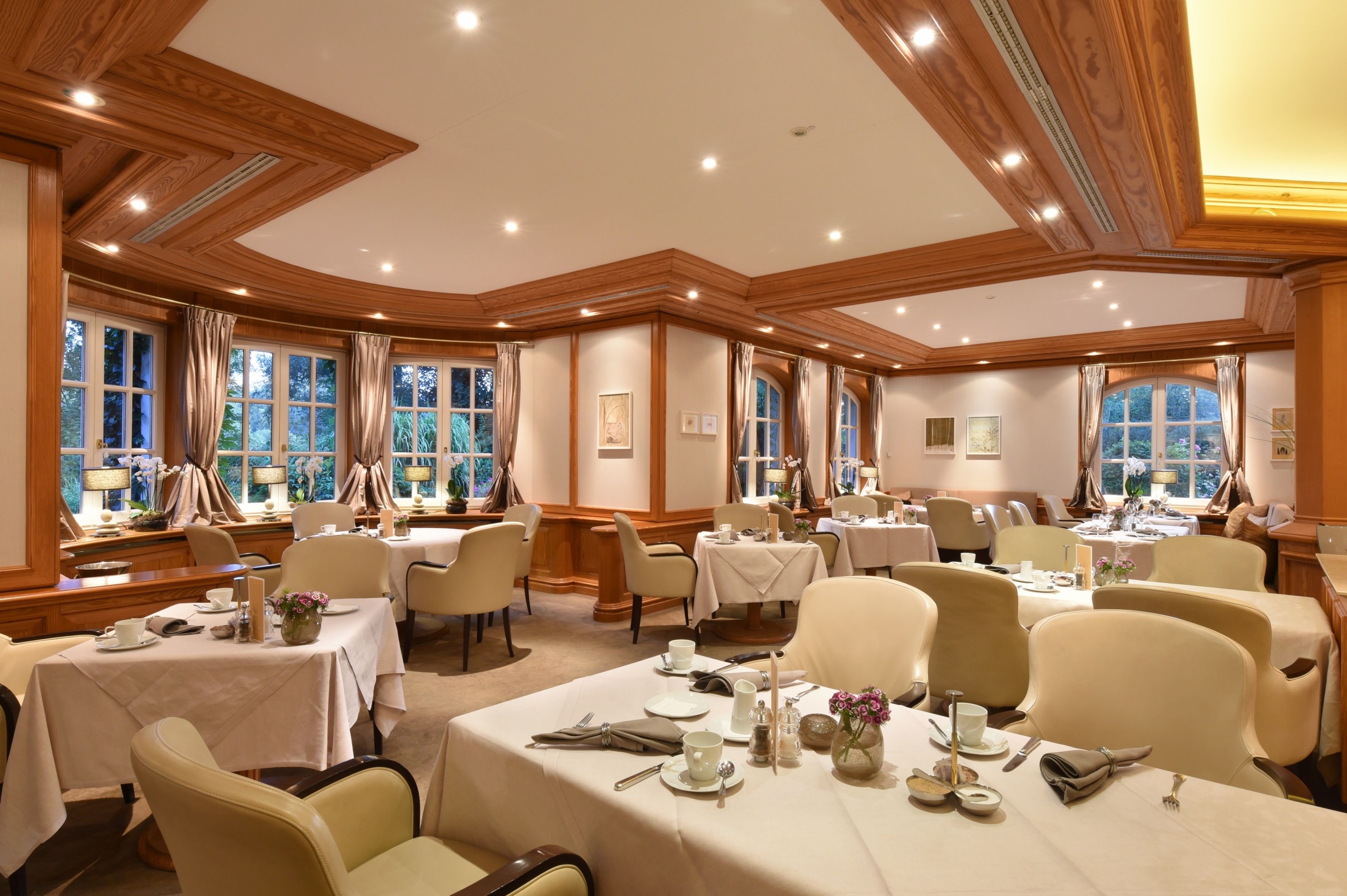 A dining area with multiple tables set for service, large windows with curtains, and a wooden ceiling design.