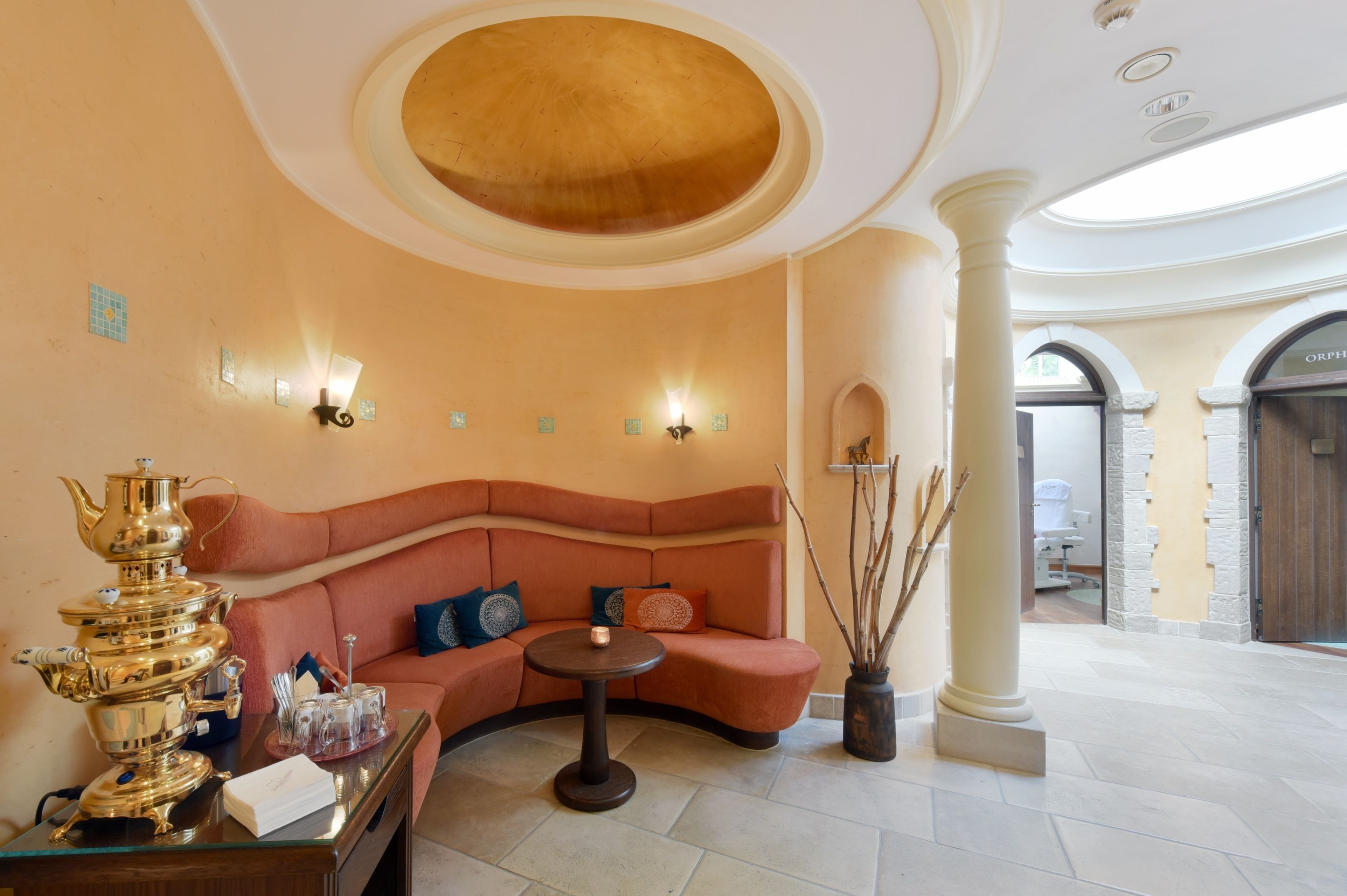 Eine luxuriöse Lobby mit einem gebogenen, orangefarbenen Sofa, einem Glastisch und einem dekorativen goldenen Teeservice.