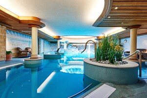 A large indoor swimming pool with a view of a waterfall, surrounded by lounge chairs and potted plants.