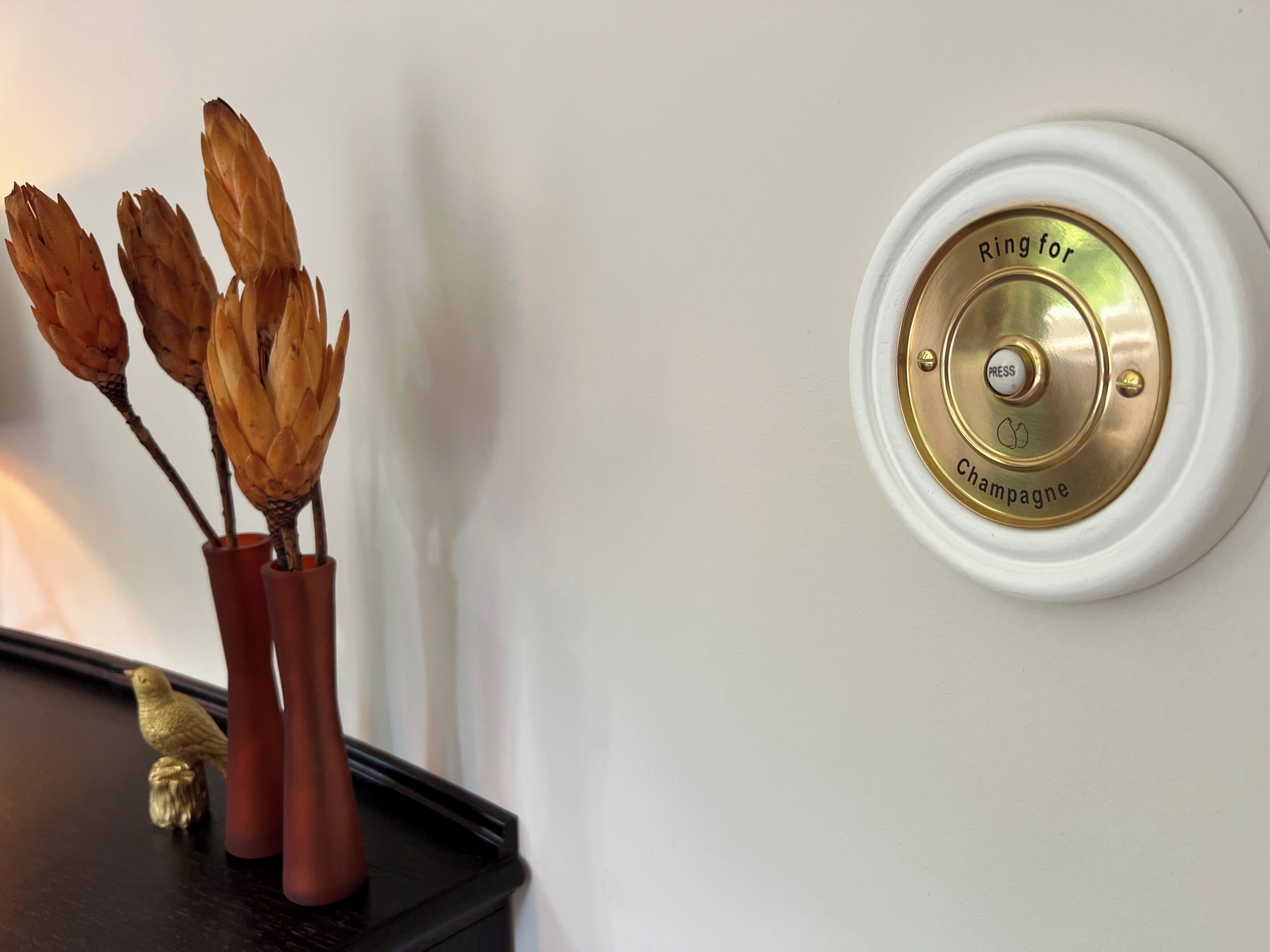 A wall-mounted device labeled "Ring for Champagne" and a vase with dried flowers.