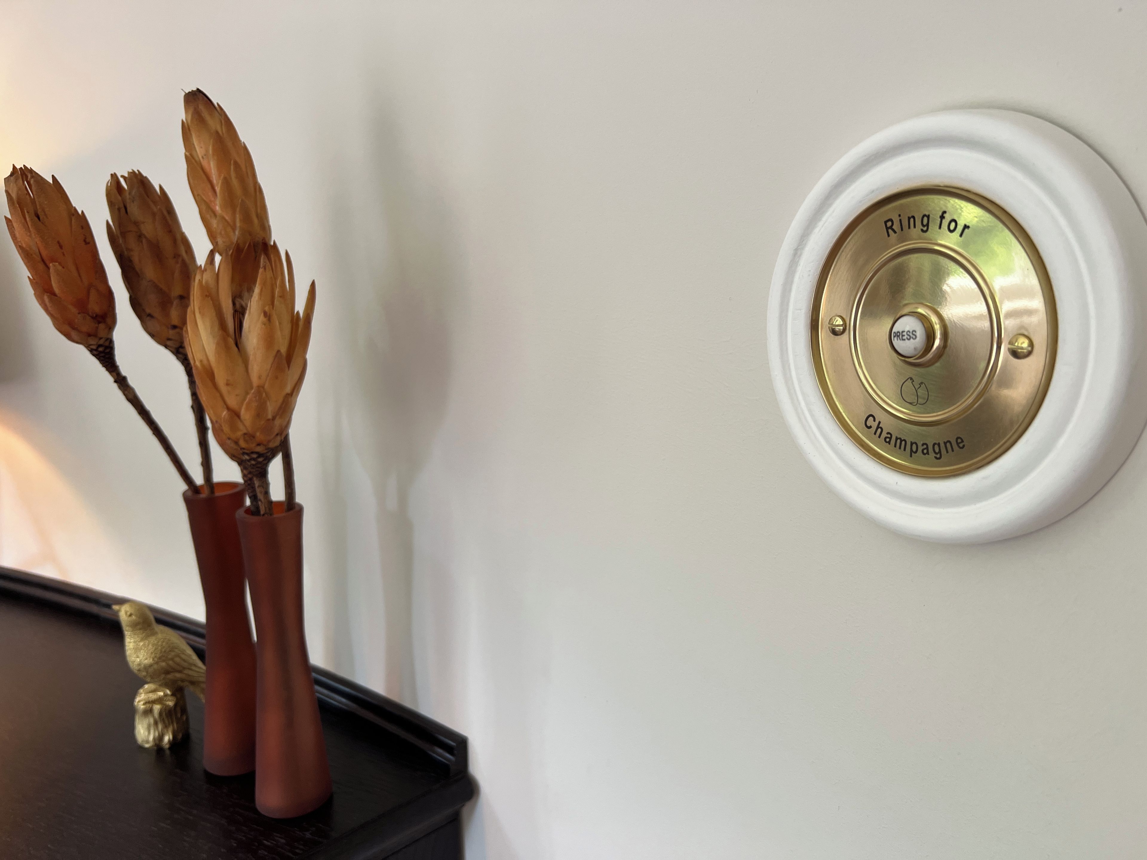 Un dispositivo fijado a la pared con la inscripción 'Ring for Champagne' y una figurita de pájaro sobre una superficie oscura.