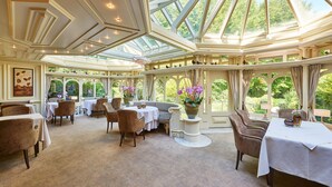 Une véranda dotée d’un haut plafond, de nombreuses tables et chaises, et de grandes fenêtres offrant une vue sur la verdure.
