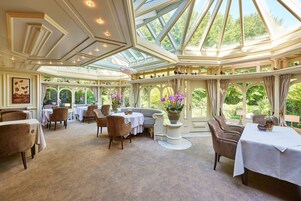 A conservatory with a high ceiling, multiple tables and chairs, and large windows offering a view of greenery.
