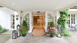 L’entrée d’un hôtel, avec une allée dallée de pierres, un escalier en bois et un espace extérieur abrité, aménagé avec des chaises et des tables.
