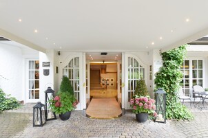 A hotel entrance with a stone-paved driveway, a wooden staircase, and a covered outdoor seating area with chairs and tables.