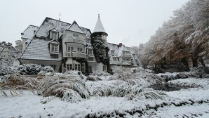 Une grande maison recouverte de neige, avec une tour imposante, entourée d’arbres et d’arbustes chargés de neige.