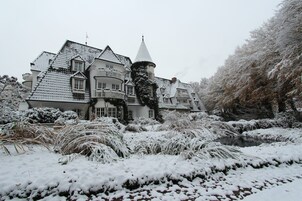 Una casa grande cubierta de nieve, con una torre destacada, rodeada de árboles y arbustos cargados de nieve.