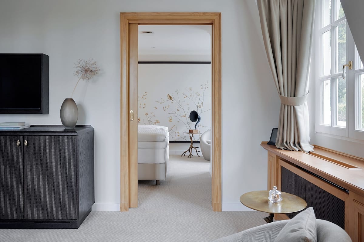 A modern hotel room with a large bed, a small round table, a TV mounted on the wall, and a view of the outdoors through a window with curtains.