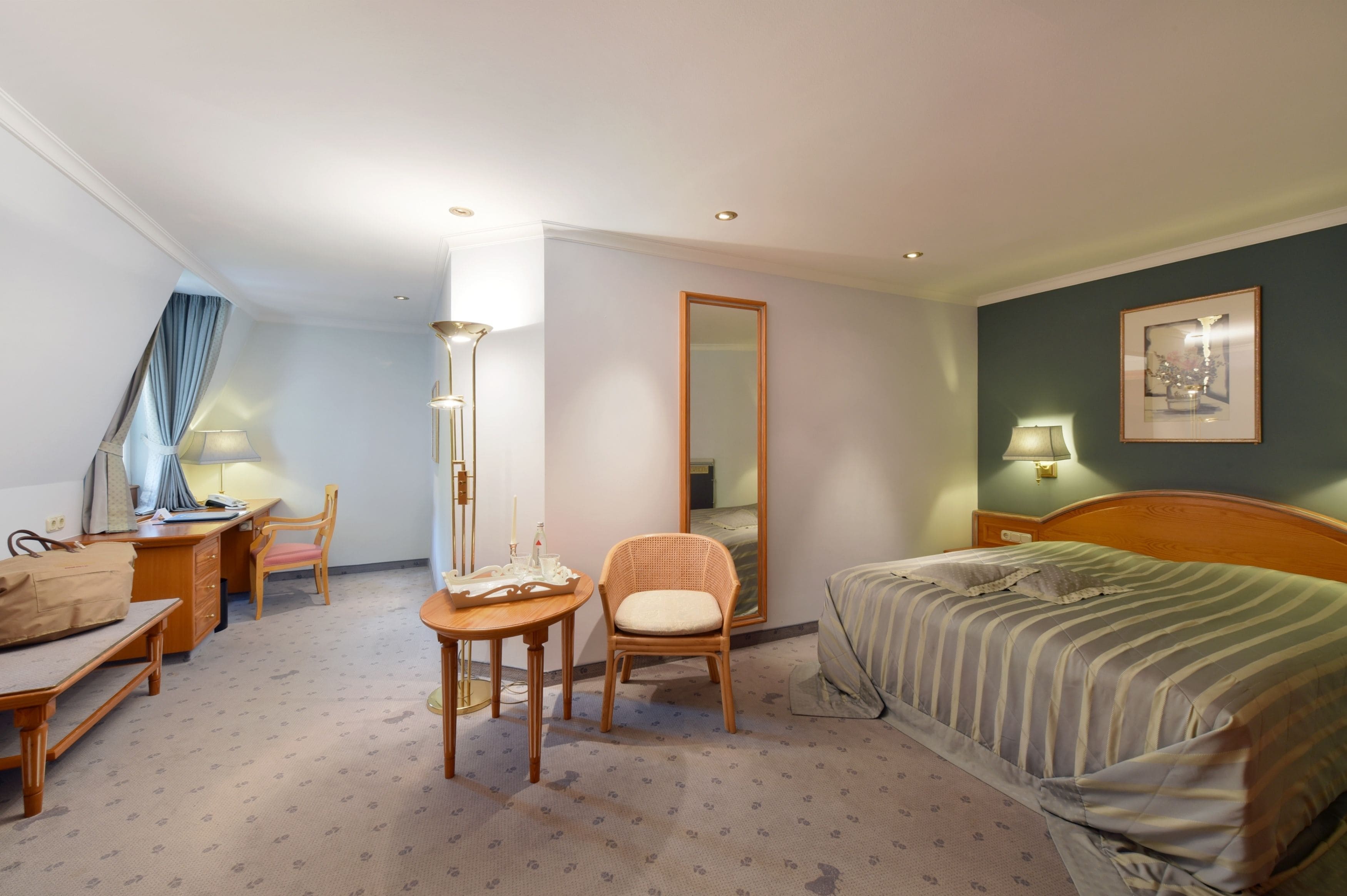 Una habitación de hotel con cama, escritorio, silla, una mesita, espejo y lámpara.