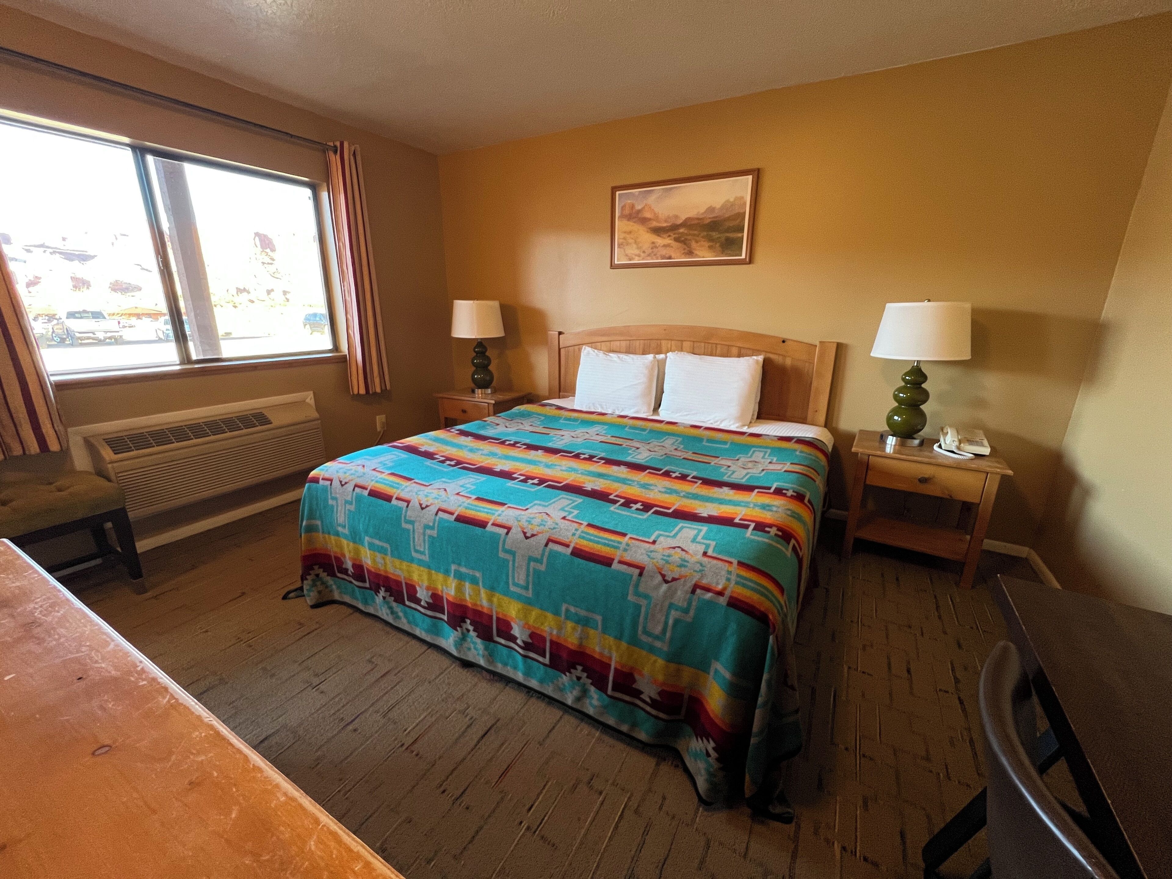 A hotel room with a large bed, two bedside tables, a window with curtains, a radiator, and a painting on the wall.