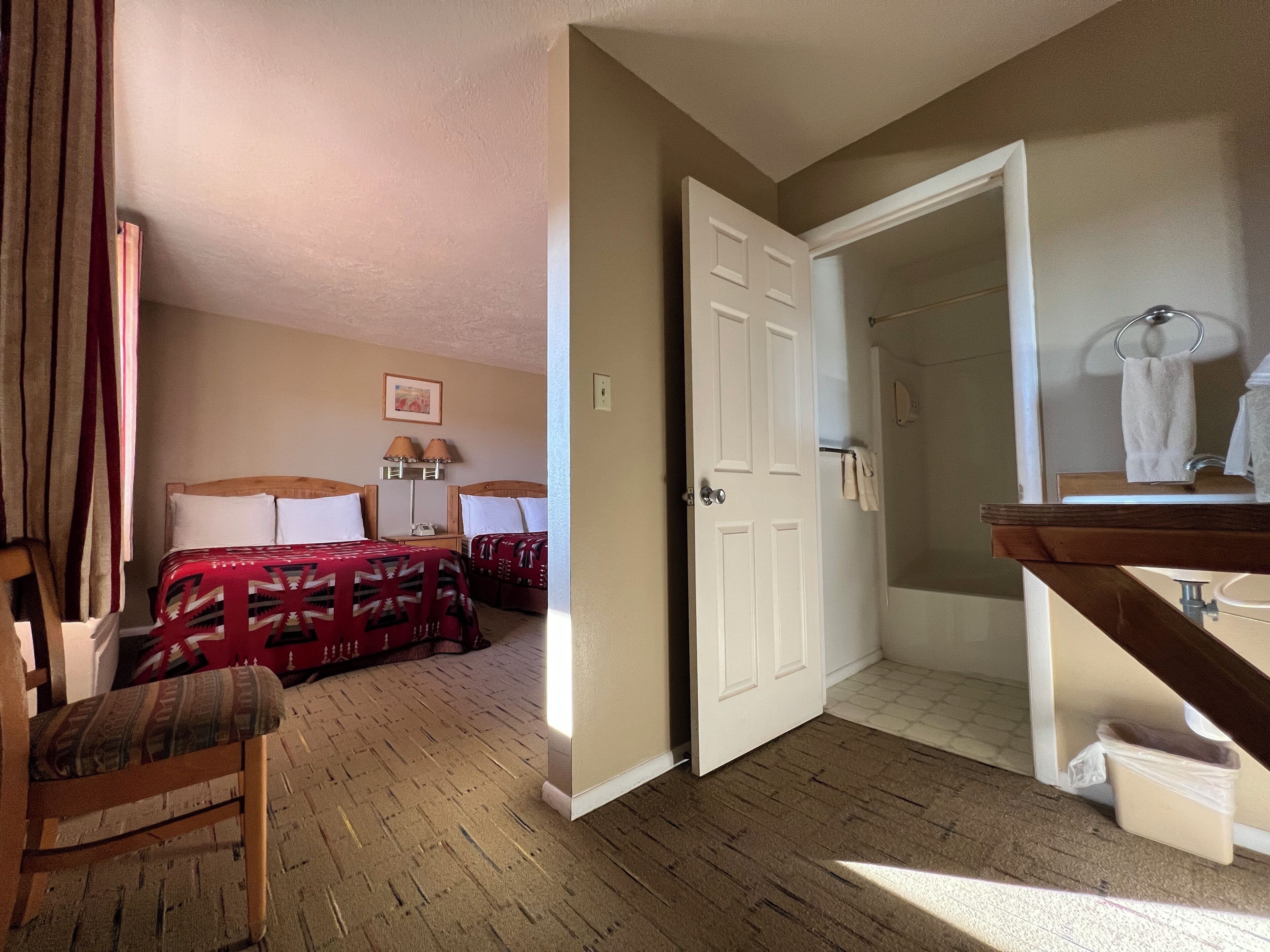 Quarto de hotel com cama, uma cadeira, escrivaninha com luminária, banheiro com pia e espelho, e porta que dá para outro cômodo.