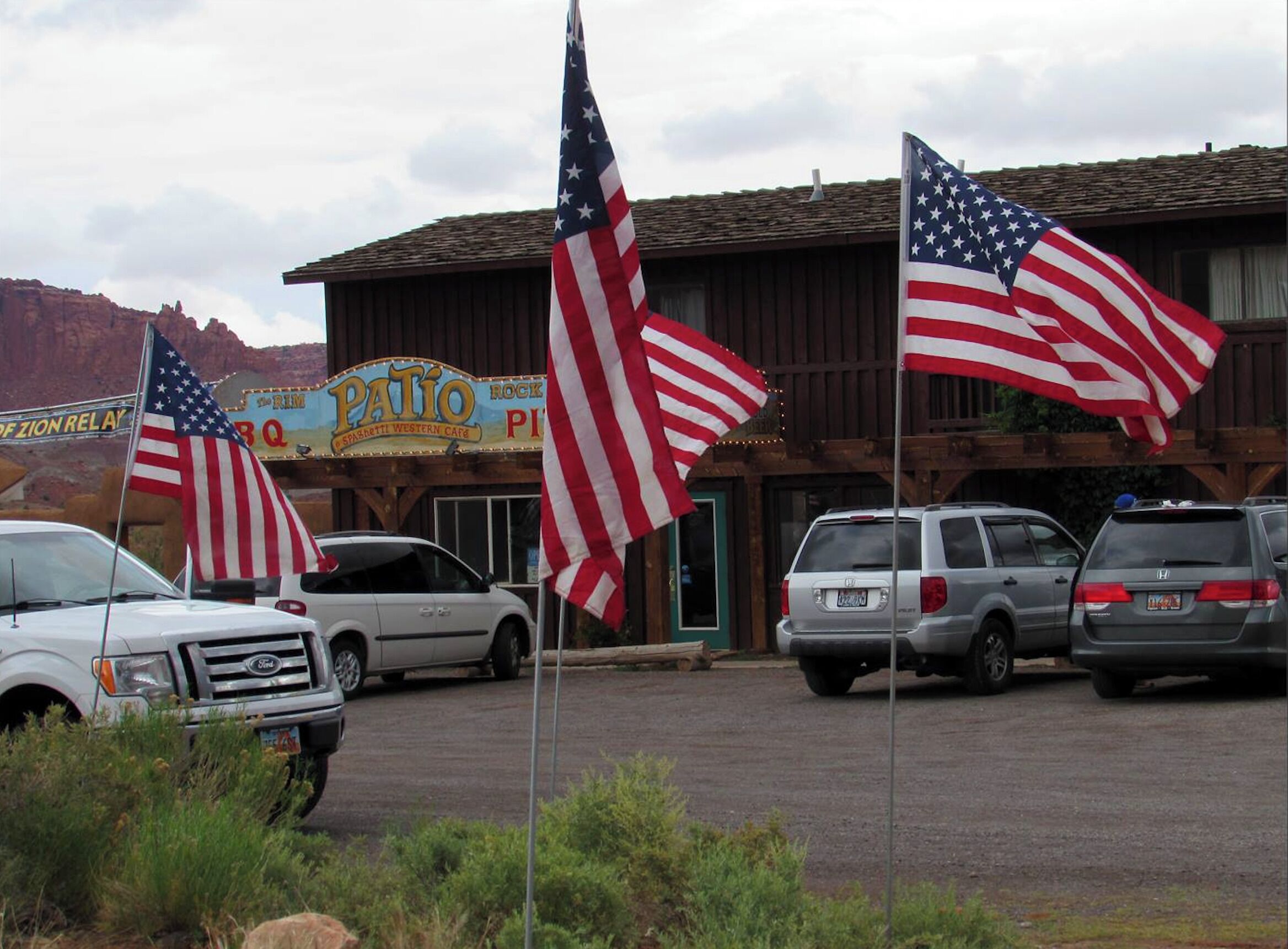 A parking lot with several cars and American flags in front of a building with a sign that reads "Patio Rock Pit BBQ".