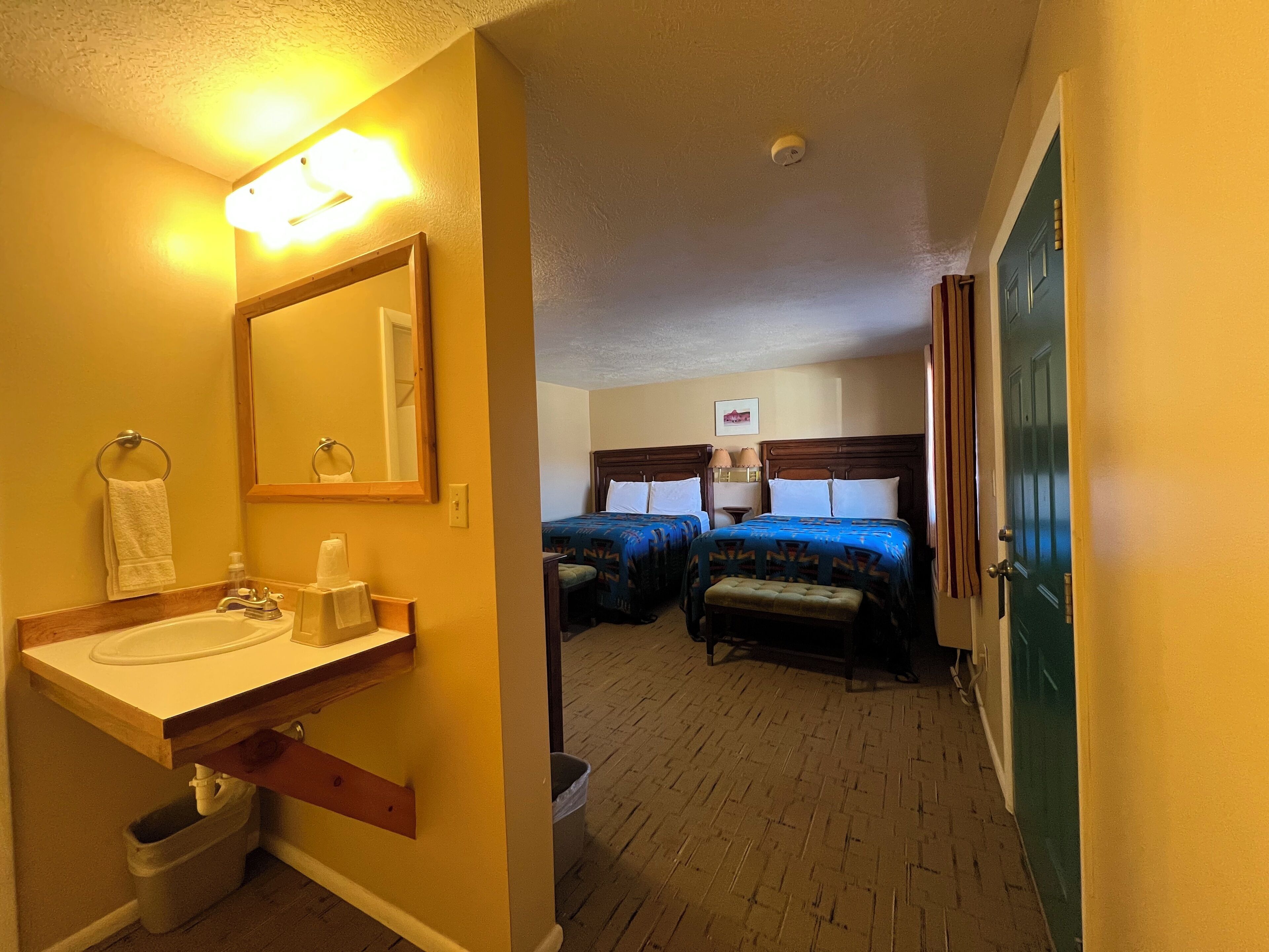 Une chambre d’hôtel avec salle de bain, deux lits, un banc en bois et une poubelle.