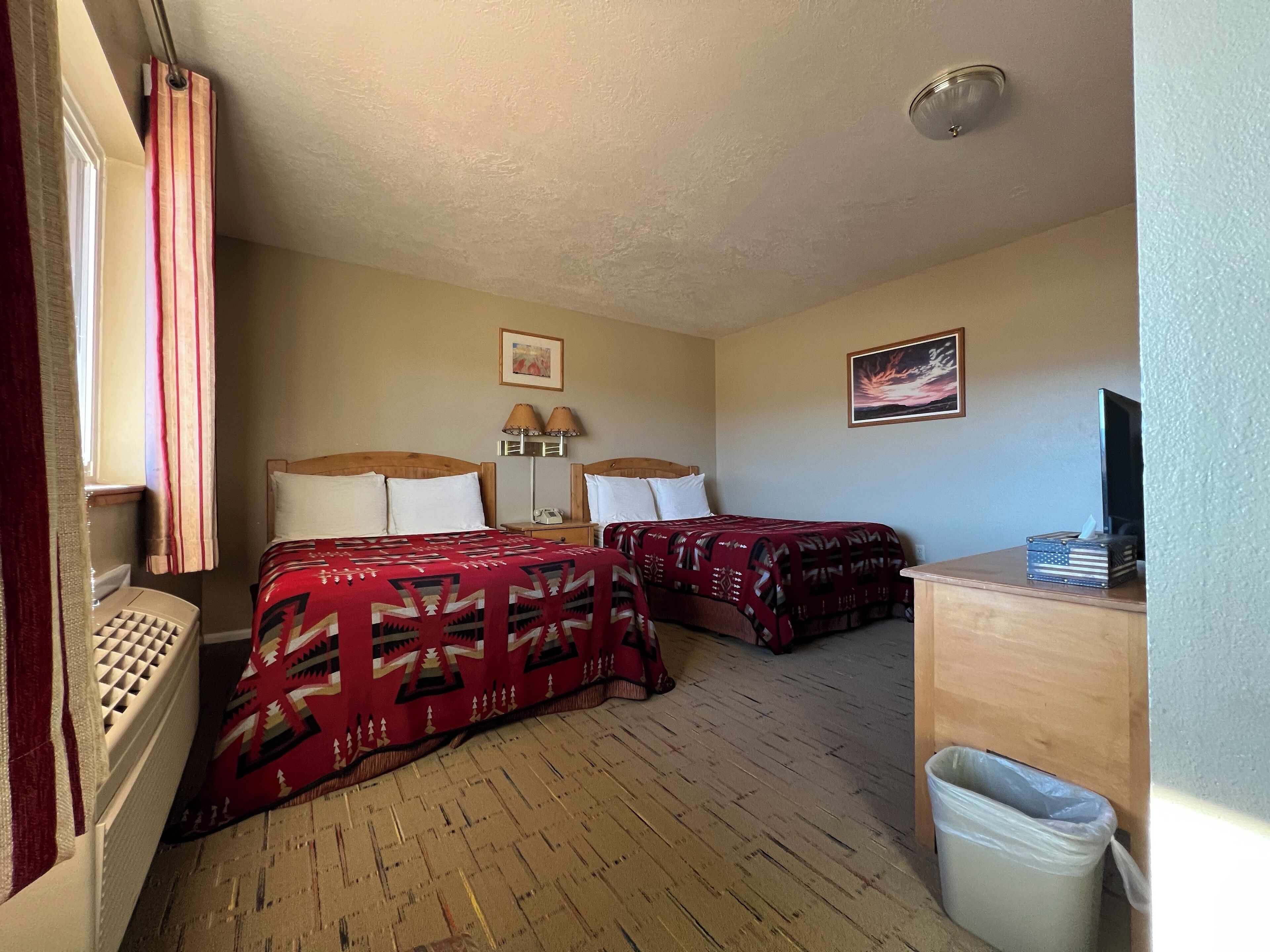 A hotel room with two beds, a desk, a trash bin, and a television.