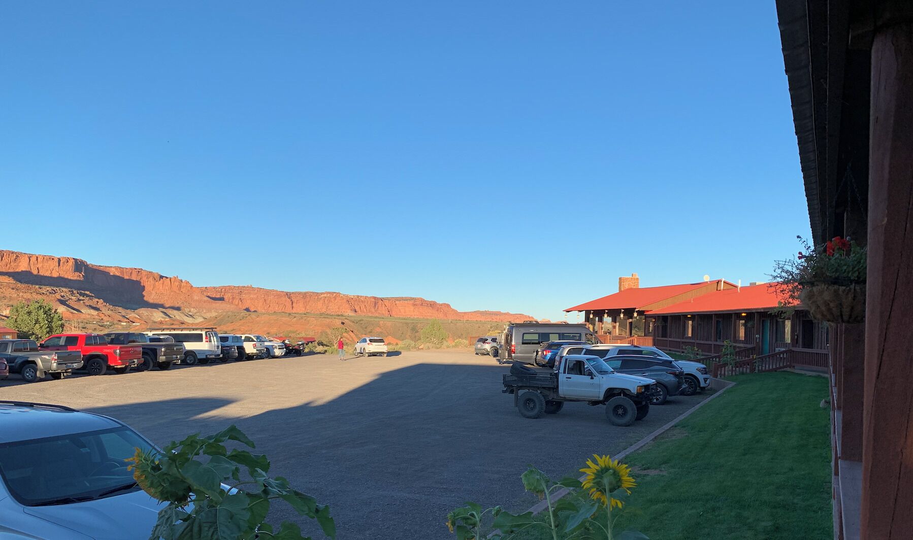 A parking lot with various vehicles, a building with a red roof, and a mountainous backdrop.