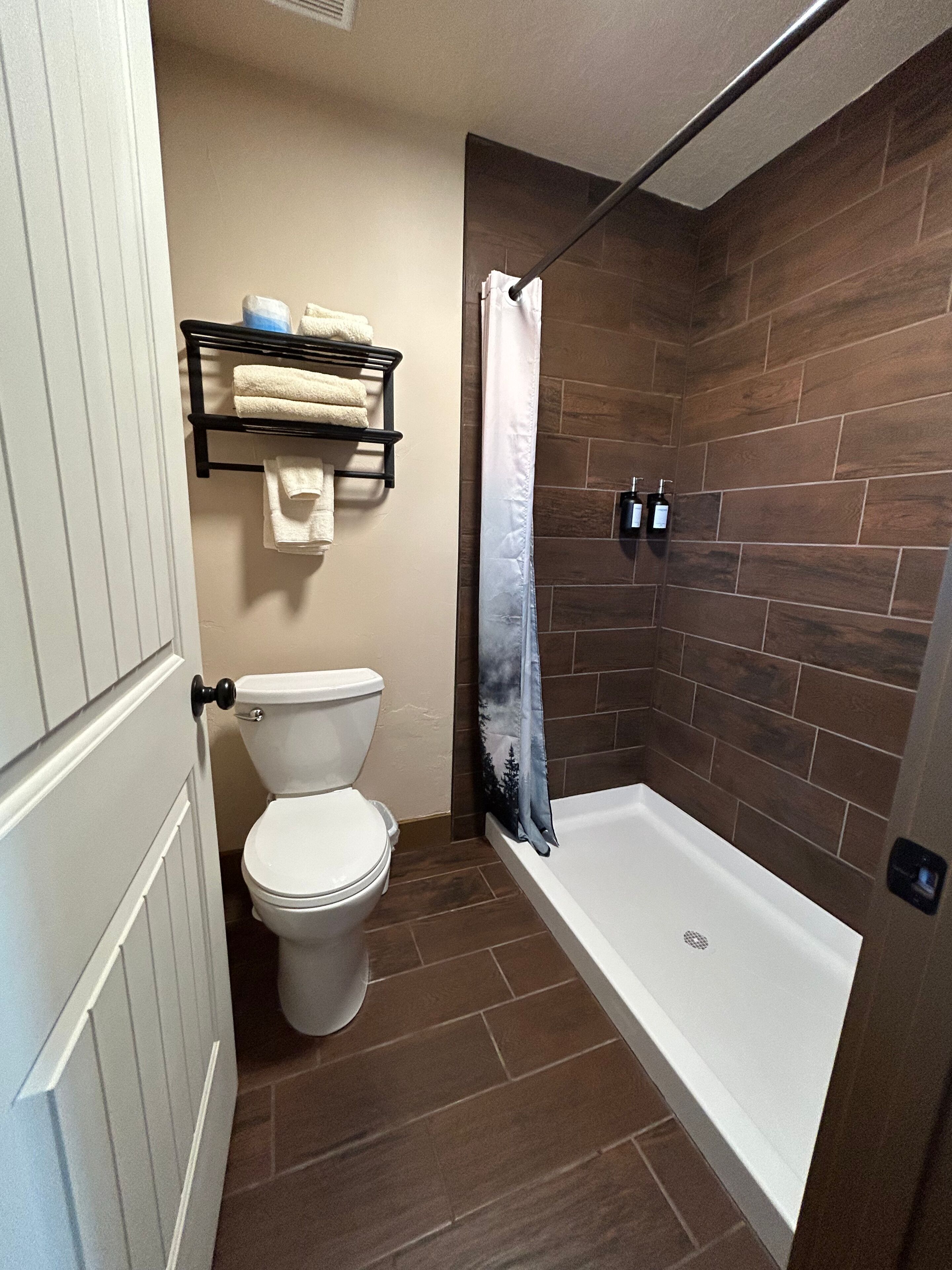 Une salle de bain avec des toilettes blanches, une douche avec un rideau, un porte-serviettes et une bouteille de shampoing.