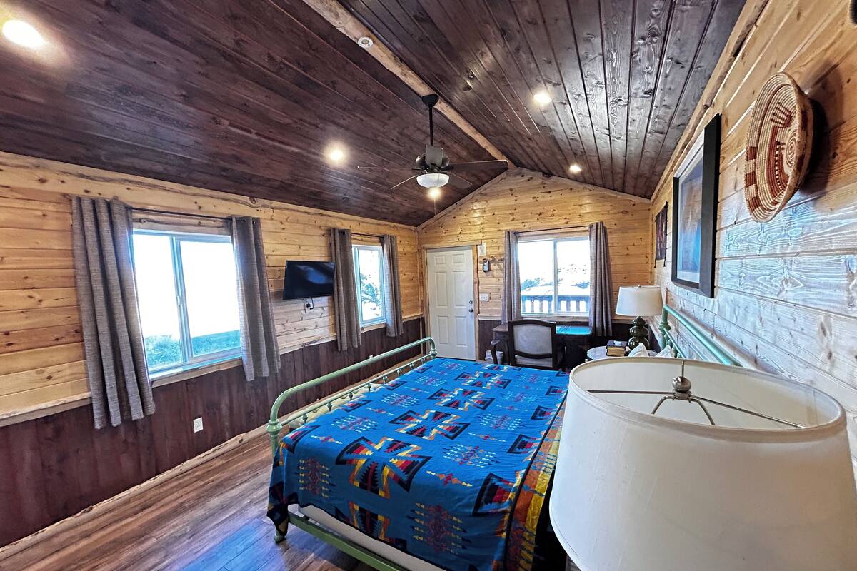 A cozy room with a wooden ceiling, a bed with a colorful quilt, a desk with a chair, and a TV mounted on the wall.
