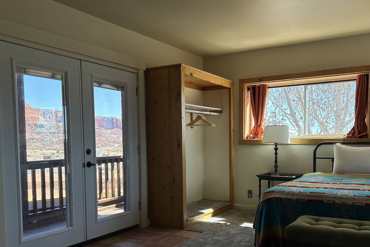 A bedroom with a bed, a nightstand, a lamp, a window with curtains, a wooden bench, and a door leading to an outdoor area.