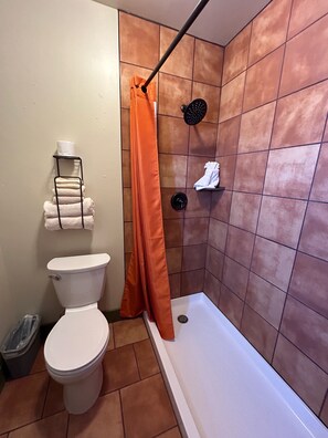 A bathroom with a toilet, a shower with an orange shower curtain, and a towel rack with folded towels.