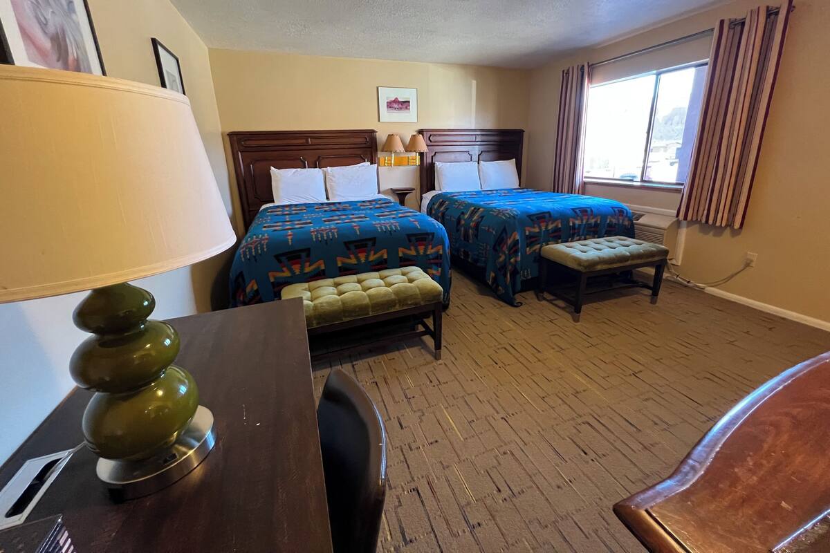 A hotel room with two beds, a desk, a lamp, and a window with curtains.