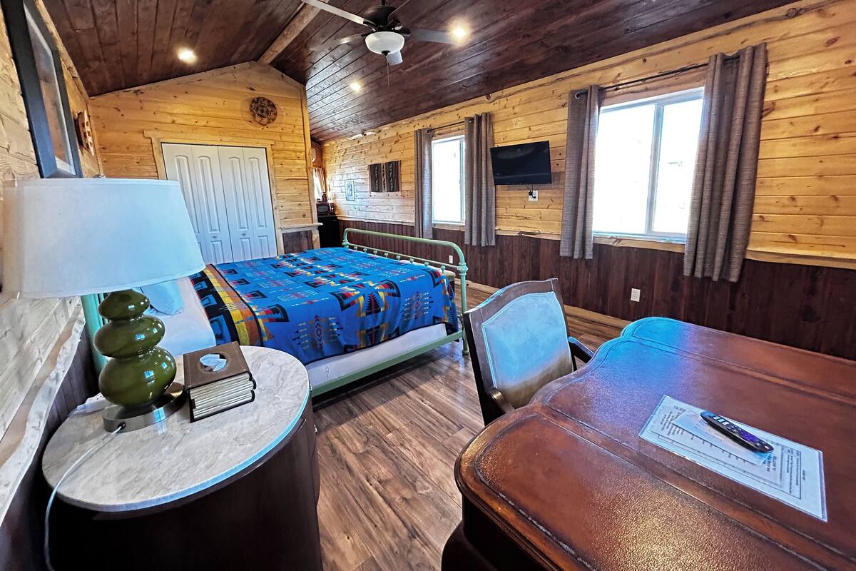 A wooden cabin room with a bed, a table, a lamp, and a television on the wall.