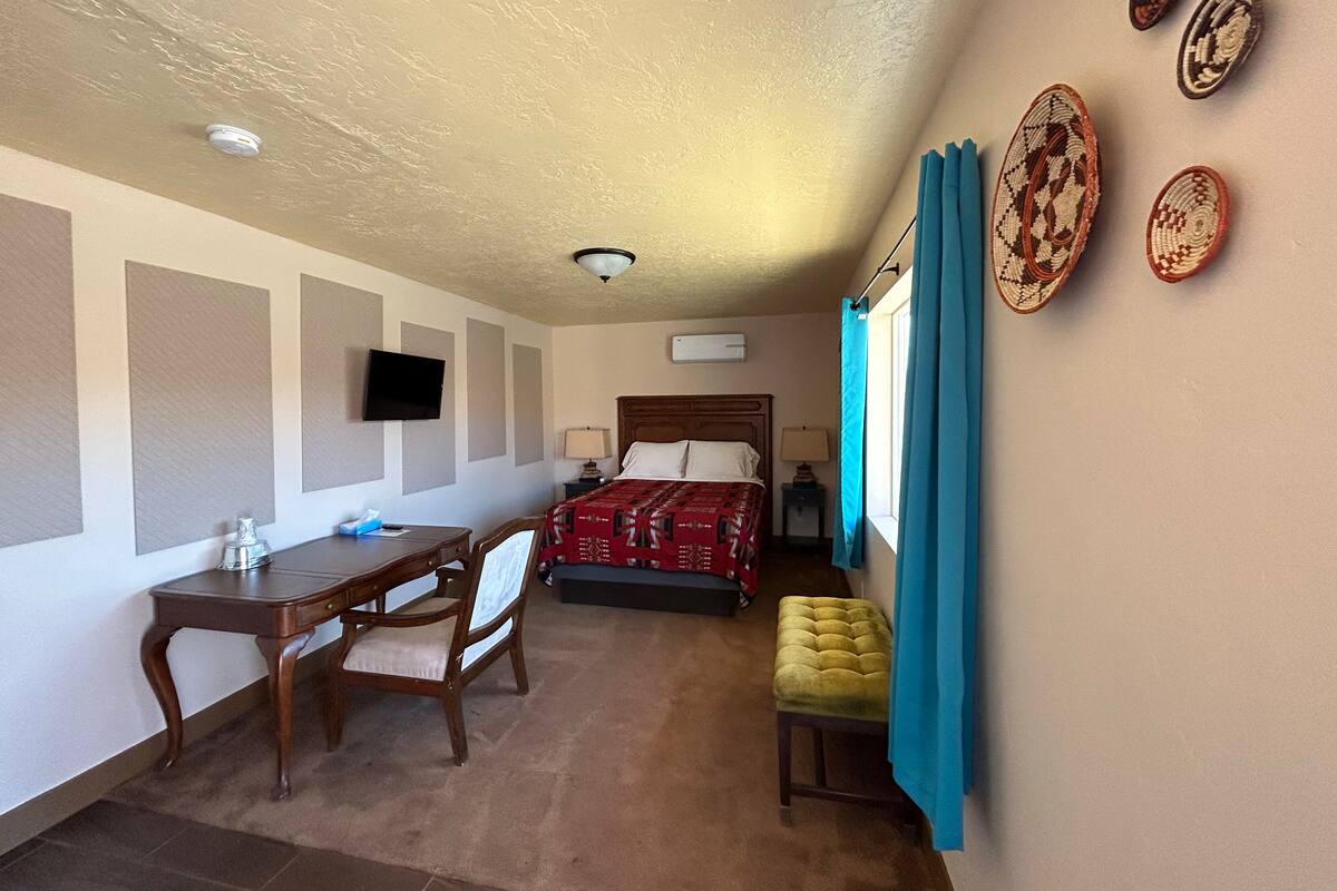 A hotel room with a bed, a desk, a chair, a small table, a TV, and a window with curtains.