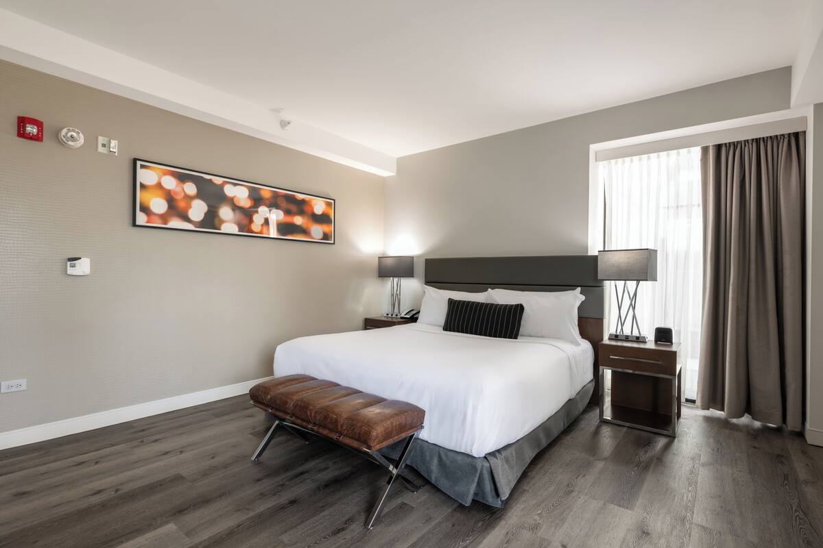 A modern hotel room with a large bed, a bench, a nightstand, a lamp, and a window with curtains.