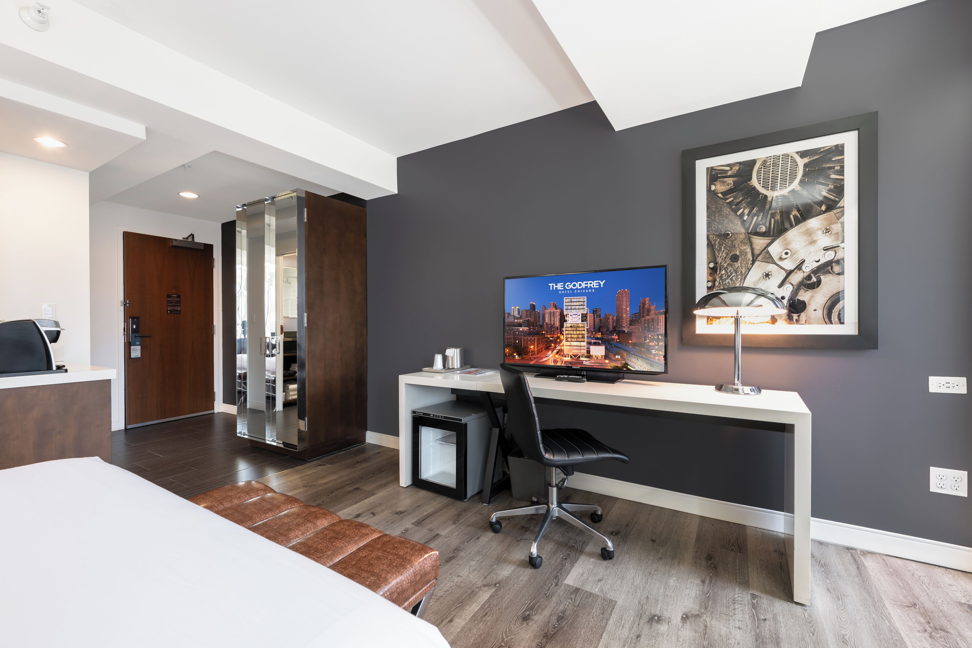 A modern hotel room with a large bed, a desk with a computer, a television displaying 'The Godfrey', and a framed abstract artwork on the wall.
