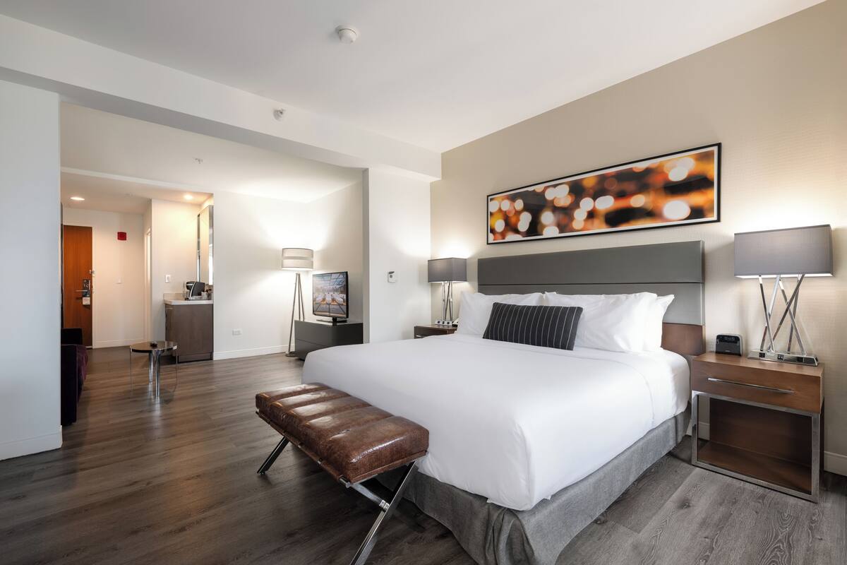 A modern hotel room with a large bed, bedside tables, a bench, and a painting on the wall.