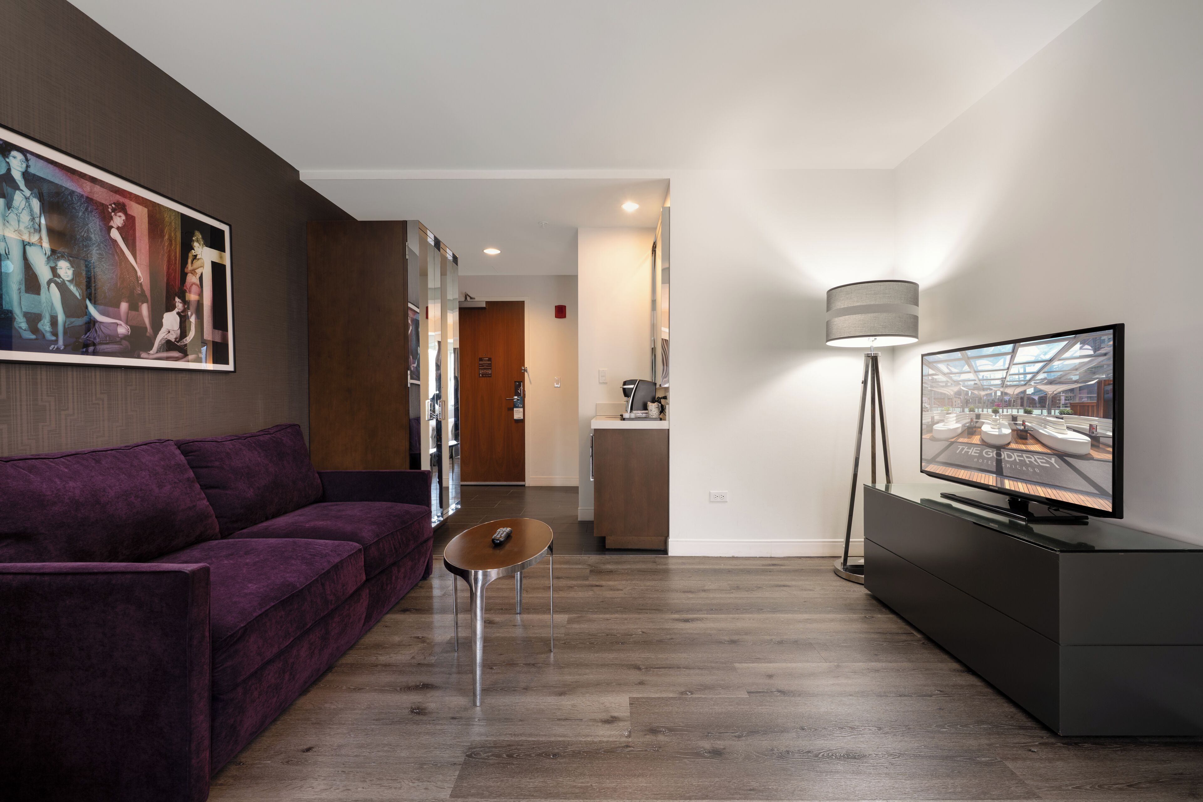 A modern living room with a purple sofa, a small table, a TV on a stand, and a painting on the wall.