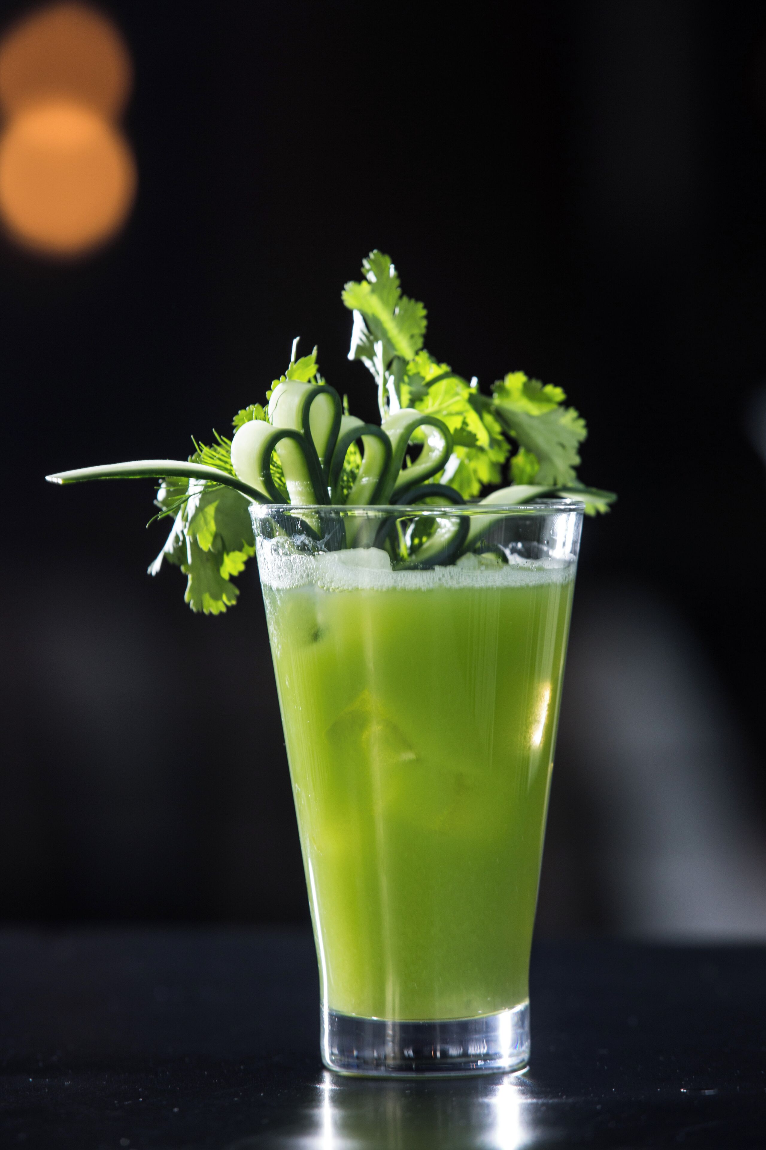 Un vaso alto lleno de una bebida verde, adornado con hierbas y una hoja verde, sobre una superficie reflectante.